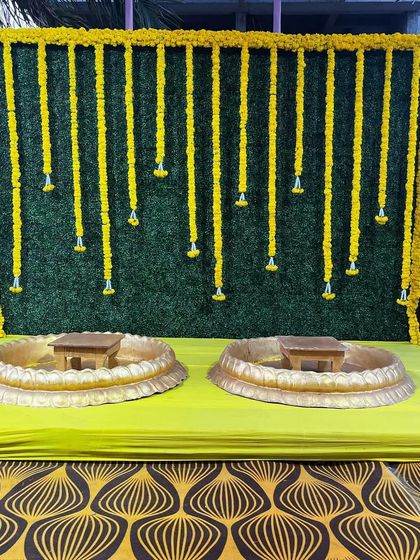 A simple yet classic Haldi backdrop. This setup uses a green foliage wall with hanging marigold strings and two traditional urlis, perfect for a small and intimate ceremony.