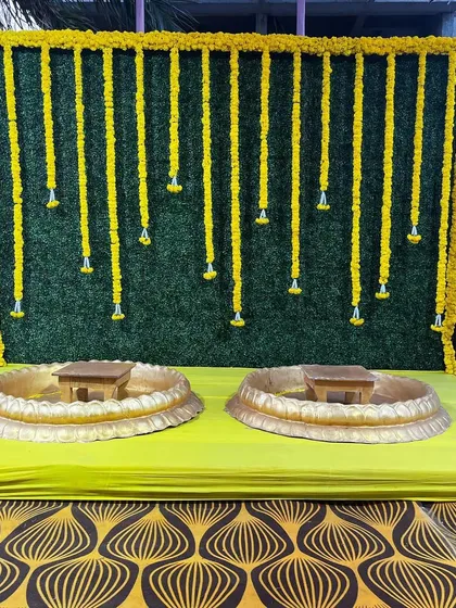 A simple yet classic Haldi backdrop. This setup uses a green foliage wall with hanging marigold strings and two traditional urlis, perfect for a small and intimate ceremony.