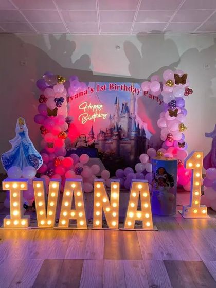 A grand Disney Princess theme for a first birthday party. This setup features a castle backdrop, character cutouts of Ariel and Cinderella, and large light-up letters spelling the birthday girl's name.