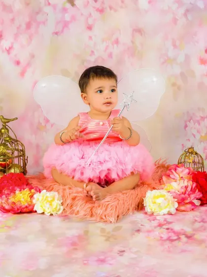 A baby girl dressed as a fairy in a pink tutu, complete with wings and a wand, sits in a dreamy floral setting.