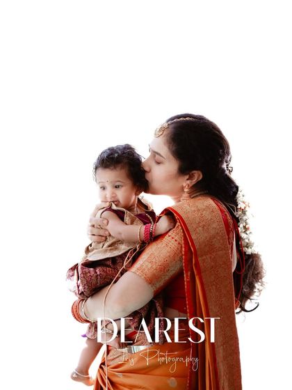 A tender moment between a mother and her baby during a naming ceremony. The mother, dressed in a beautiful orange saree, gently kisses her child's head in this high-key portrait.
