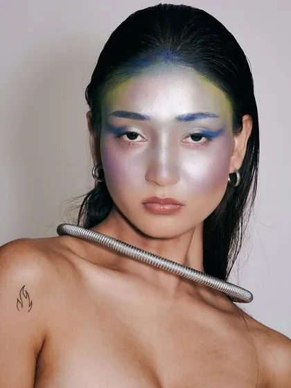 A futuristic portrait with iridescent, multi-toned makeup creating an alien-like contour on the face. The hair is kept simple and sleek to not distract from the colorful, metallic skin effect.