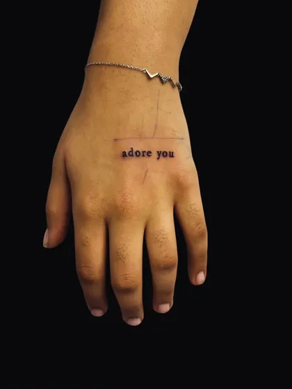 The phrase 'adore you' in a simple typewriter font on the hand. This minimalist script tattoo is a sweet and constant reminder.