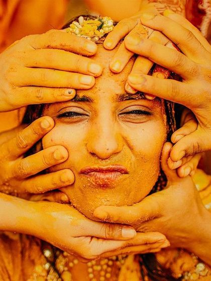 A classic Haldi moment, with the bride's face being lovingly covered in turmeric paste by her family. Her scrunched-up expression captures the fun and messy reality of this vibrant tradition.