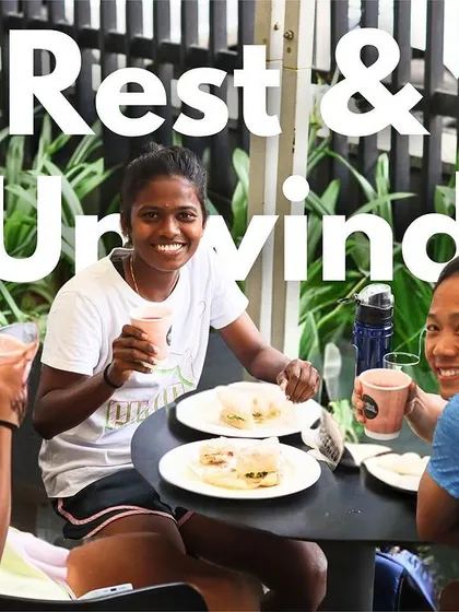 Rest and Unwind. Three athletes, including Bala Devi, smile and relax together. This image captures the supportive and friendly atmosphere we foster among our athletes.
