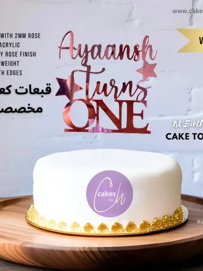 A mockup of the "Ayaansh Turns ONE" topper in rose gold on a simple white cake, showcasing a clean and modern aesthetic.