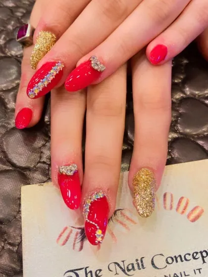 An extravagant set of red and gold almond nails. The design is packed with detail, including gold glitter, multi-colored rhinestones, and 3D bow embellishments.
