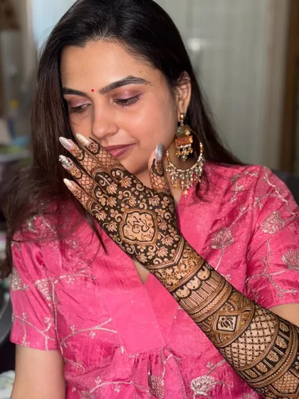 A beautiful bride showing off her intricate back-of-hand mehendi. The design features a central motif with initials, surrounded by delicate floral patterns.