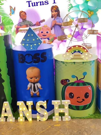 A close-up of a Cocomelon and Boss Baby-themed setup, showing the custom-printed plinths and 'ANSH' marquee letters.