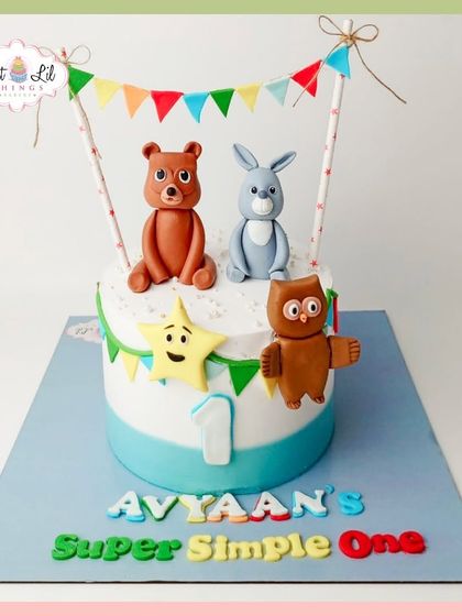 A "Super Simple" first birthday cake featuring cute woodland creatures like a bear, bunny, and owl. The colorful bunting adds a festive touch.