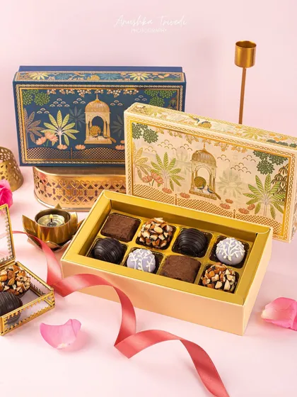 An elegant gift box of assorted chocolates, styled with delicate rose petals and brass diyas. The composition is designed to look both luxurious and inviting.