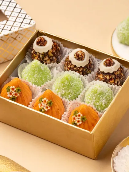 A close-up of a golden gift box filled with an assortment of colorful Indian sweets. The tight framing emphasizes the textures and artistry of each piece, from the nutty ladoos to the coconut-covered modaks.
