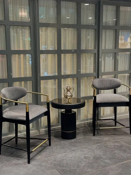 A sophisticated pairing from our exhibition: two of our high-back chairs with dark frames and grey upholstery, flanking a sleek black and gold side table.