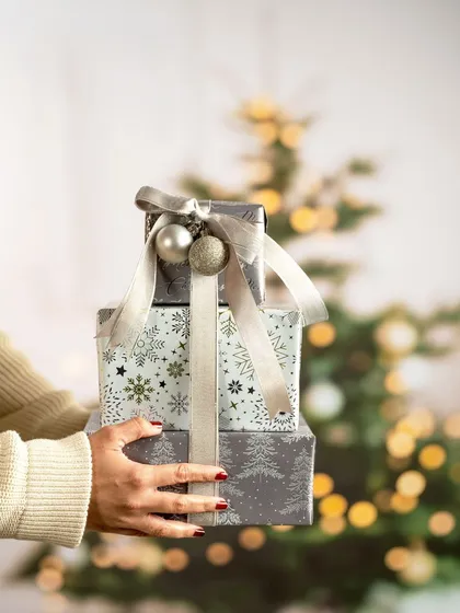A lifestyle shot of hands holding a stack of beautifully wrapped Christmas presents. The blurred Christmas tree in the background creates a festive and heartwarming mood, perfect for a holiday campaign.