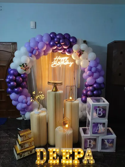 An elegant home birthday setup with a purple and white balloon arch, a warm 'Happy Birthday' neon sign, and custom light-up name display.