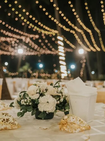 An elegant table setting for a white-themed wedding reception. The simple floral centerpiece and personalized shell place cards are set against a beautiful backdrop of bokeh fairy lights.