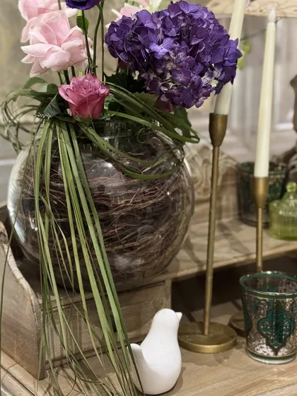 A floral arrangement in a glass bowl filled with natural twigs, paired with a small white bird figurine, showcasing an artistic and organic approach to centerpieces.