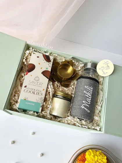 A personalized Diwali hamper in a beautiful pastel green box. It includes a custom-named bottle, coffee-filled cookies, a scented candle, and a diya, making it a thoughtful and complete gift.