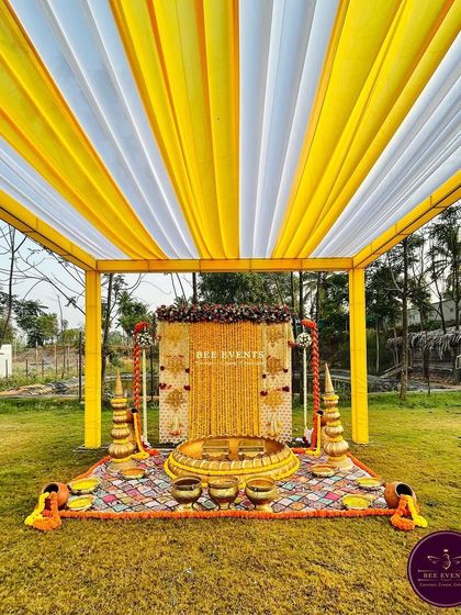 A complete view of an outdoor Haldi setup. The yellow and white draped canopy, the marigold backdrop, and the traditional brass vessels create a picture-perfect scene for the ceremony.
