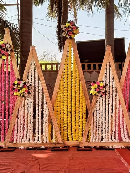 A unique and modern Haldi backdrop featuring triangular wooden frames adorned with pink, white, and yellow flower garlands.