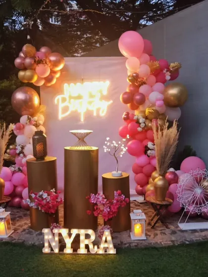 A beautiful pink and gold themed birthday party. The decor includes a custom backdrop with a neon sign, metallic gold pedestals, a lush balloon garland, and personalized light-up letters.