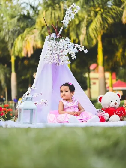 A wider view of the pink-themed outdoor picnic setup. It shows the placement of the teepee, 'ONE' sign, balloons, and teddy bear to create a full and balanced scene.
