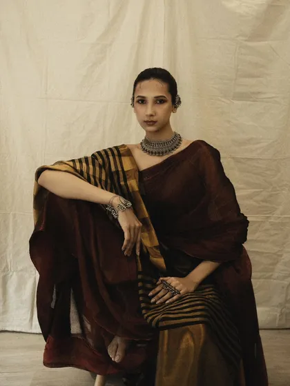 A full-length seated portrait that displays the entire saree, including the contrasting striped pallu. The pose is grounded and elegant, with a timeless quality.