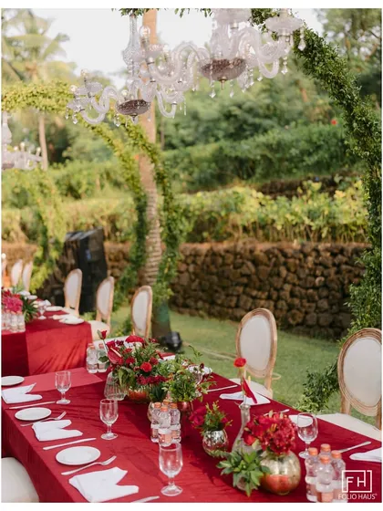 An elegant outdoor dinner setup with a bold red theme. The long tables are adorned with red linens, floral centerpieces, and crystal chandeliers hanging from the trees.