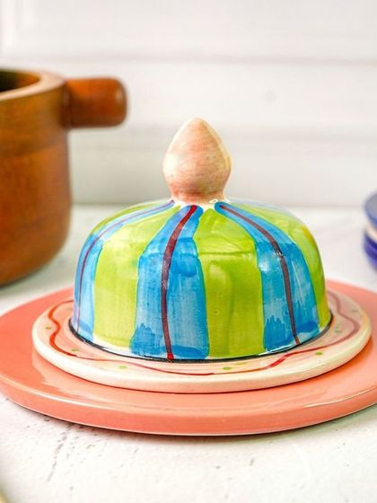 A colorful, hand-painted butter dish styled in a kitchen scene with a wooden bowl of apples. This shows the product in a natural, homey setting.