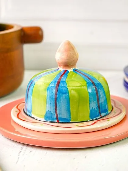 A colorful, hand-painted butter dish styled in a kitchen scene with a wooden bowl of apples. This shows the product in a natural, homey setting.