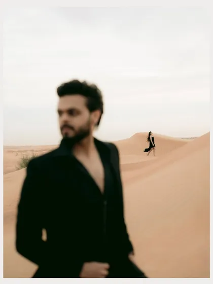 An artistic, unconventional portrait in the Dubai desert. The groom is in soft focus in the foreground while the bride stands sharply in the distance, creating a sense of depth and story.