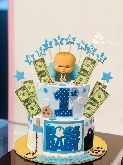 A two-tier 'Boss Baby' themed cake for a first birthday. This fun design is complete with edible money, a briefcase, and a milk bottle to celebrate the little boss in the family.