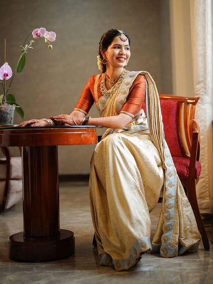 A beautiful, naturally lit portrait of a bride in a classic off-white and rust-orange silk saree. The makeup is kept soft and natural to enhance her features, creating a look of pure elegance.