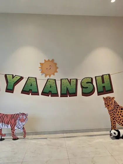 A cute and simple jungle-themed backdrop for Ayaansh's first birthday. The setup includes tree and animal cutouts like a tiger, leopard, and bear, with a personalized name banner.