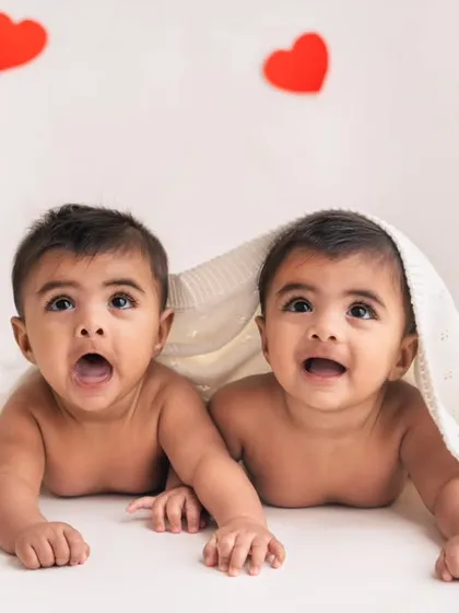 Double the love! These twin brothers are peeking out from under a blanket during their half-birthday photoshoot, set against a simple backdrop with hearts.