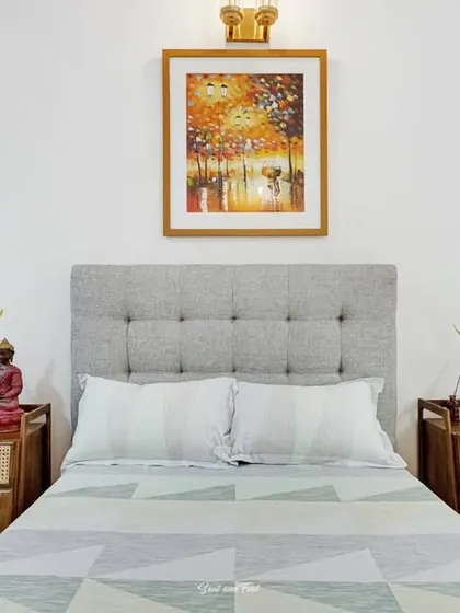 A cozy bedroom with a tufted grey headboard and light wood furniture. A colorful painting adds a splash of vibrancy to the otherwise calm and neutral space.