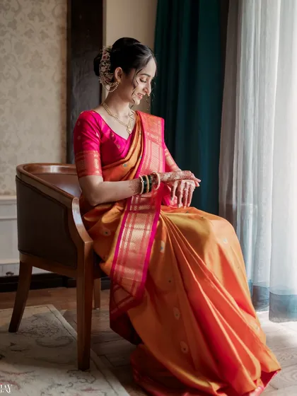 A serene, seated portrait of the bride by a window. The soft, directional light highlights the graceful drape of her orange saree and the traditional green bangles, a key part of the Marathi bridal look.