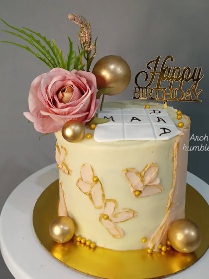 A moist chocolate cake decorated with a pink rose, golden spheres, and butterfly accents.