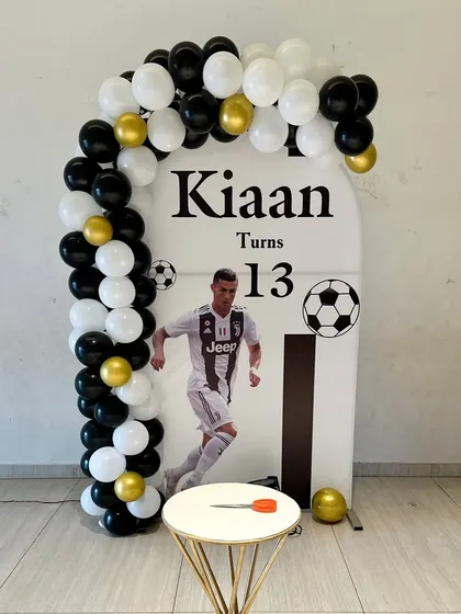 A football-themed setup for a 13th birthday. The decor features a personalized backdrop with the birthday boy's favorite player, a simple balloon arch, and a soccer ball motif.