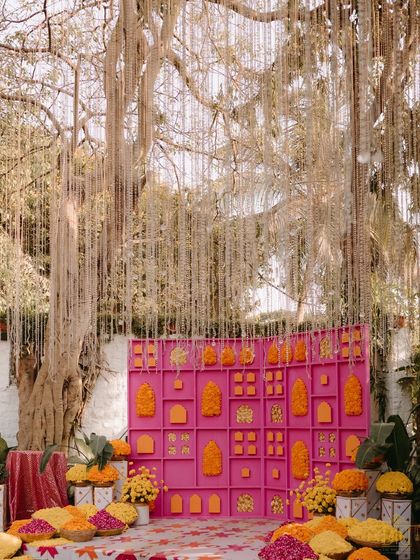 A bright pink backdrop decorated with marigolds and traditional motifs served as a perfect photo op at the Mehendi. Above, strings of pearls cascaded from a large tree, adding a touch of elegance to the vibrant setup.