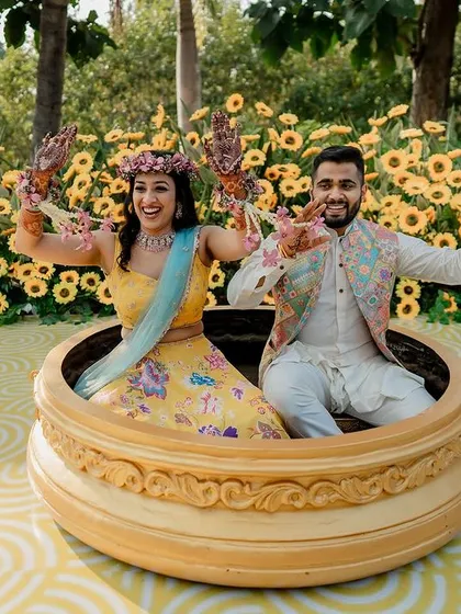 A playful shot of the couple during their Haldi ceremony, seated in a giant brass urli and holding floral kaleeras.