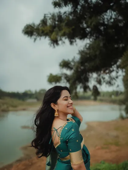 A candid, smiling shot of actress Mirnalini Ravi by the water. Her natural makeup and windswept hair create a beautiful and serene image.