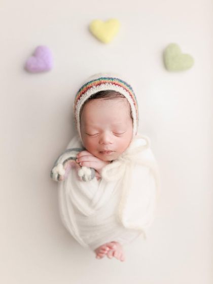 A minimal rainbow theme. The simple white wrap and rainbow-trimmed bonnet, paired with pastel felt hearts, create a subtle and sweet celebration.