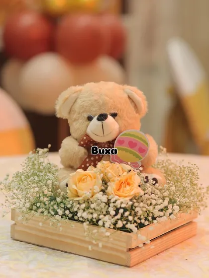 A close-up of the teddy bear centerpiece, showing the soft toy nestled among fresh roses and baby's breath.
