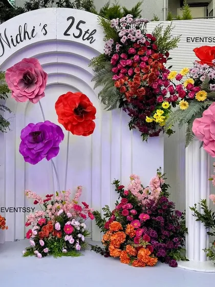 The central part of the 25th birthday backdrop, showing the custom "Nidhi's 25th" signage inside a modern arch. The arrangement of large paper flowers and fresh blooms is designed to look artistic and abundant.