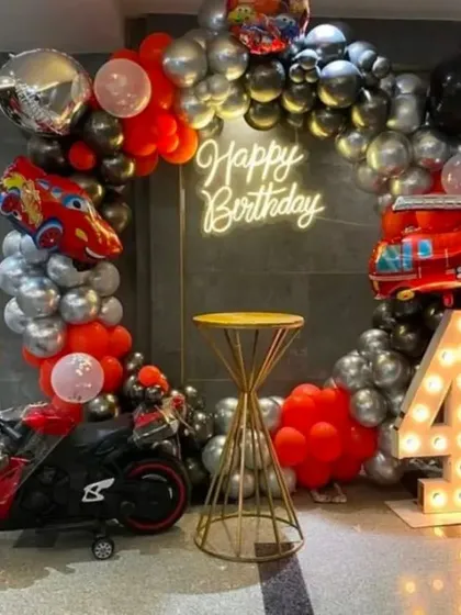 A 'Hot Wheels' and cars themed 4th birthday. The decoration includes a black, red, and silver balloon arch, car and fire truck foil balloons, a light-up number '4', and even a mini toy motorbike.