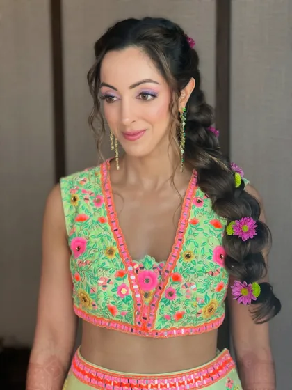 A vibrant and fun hairstyle for a mehendi ceremony. This thick, messy braid is adorned with colorful pink and green flowers to match the client's outfit.