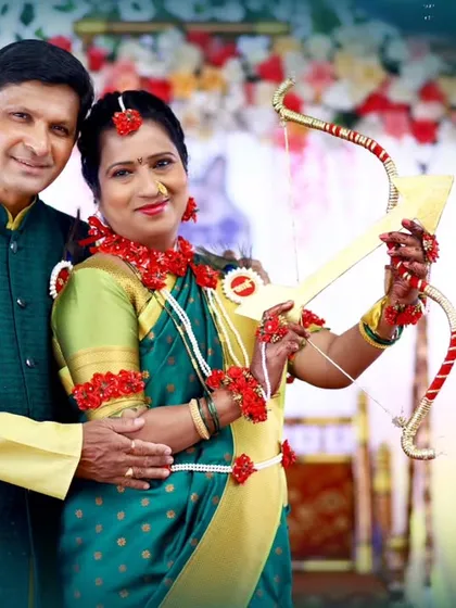A smiling couple poses with a bow and arrow prop during their Dohale Jevan, with a festive floral and character-themed backdrop behind them.