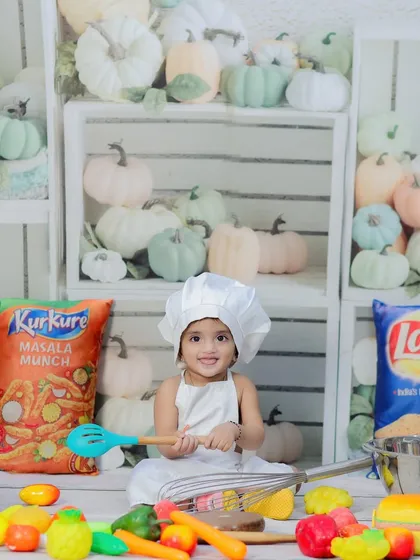 A baby girl dressed as a chef, surrounded by toy vegetables, snacks, and a wall of pumpkins. A playful and colorful setup for a foodie-in-training.