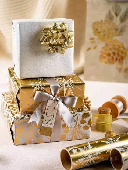 A stack of festive gifts wrapped in luxurious gold and white paper from Crown Greetings. The warm lighting and "Fa La La" tag create a classic and joyful Christmas atmosphere.